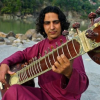 A musician in a maroon kurta plays a sitar on a quiet riverside, inviting mindful listening and nada yoga; a peaceful scene where sound, silence, and stillness help you feel present.