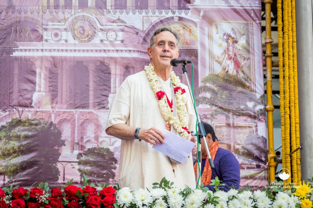 Devotional speaker wearing a white robe and floral garland addresses an audience at a temple stage, holding notes by a microphone, with an ornate temple backdrop and a row of flowers; Nectar, testing.