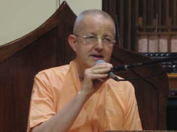 Person in orange robes speaks into a wired microphone at a lectern before a tall wooden chair in an indoor hall; image for a post on Romapada Swami guidance, testing ways to build Vaishnava ties.
