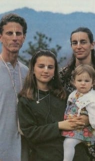 Outdoor portrait of four people before misty mountains—two adults, a teen, and a toddler—facing the camera with simple clothing, white forehead markings, and necklaces; header image for an article.
