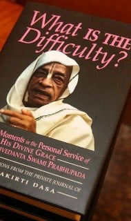 Close-up of a hardcover book titled "What is the Difficulty?", showing a portrait of an elderly spiritual teacher in white robes on the cover, resting on a table; nonfiction, spirituality articles.