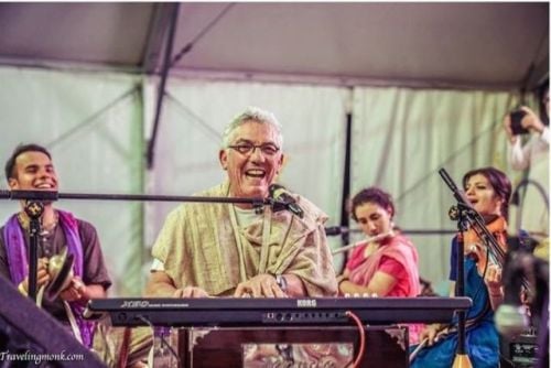 Smiling older musician in a beige shawl plays a keyboard and sings into a mic beneath a tent, surrounded by singers and a violinist; part of our Recent Media interview coverage.