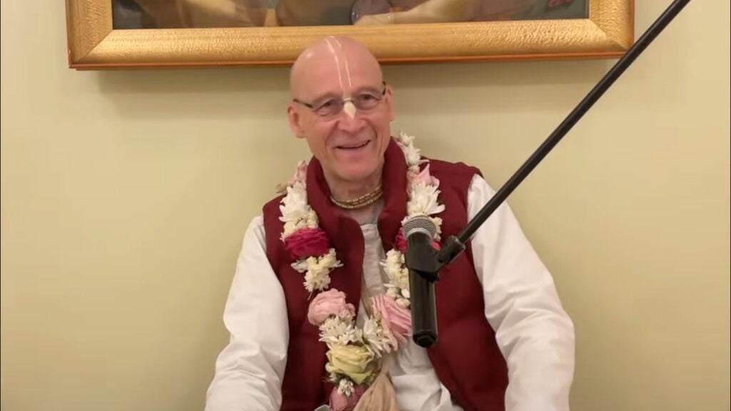 A smiling speaker wearing a flower garland sits by a microphone during a Morning Class on Srimad-Bhagavatam 10.7.19, indoors beneath a gold-framed painting; June 21, 2026, testing category.