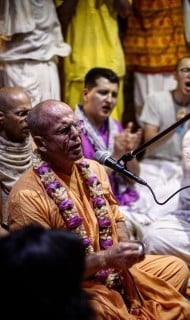 A person in orange robes with a flower garland leads call-and-response singing at a microphone while a group claps behind them in a hall, for an article on resilience, focus, and communal support.