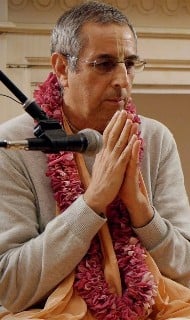 Man with glasses and a flower garland sits near a microphone, hands pressed in a prayerful gesture, addressing an indoor audience; contemplative portrait for Articles on compassion and service.