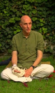 Calm garden scene: a bald adult sits cross-legged on grass before a leafy wall, wearing a green shirt and light pants, holding a small cloth bag, appearing engaged in conversation and focus.