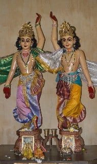 Two ornately dressed Hare Krishna deity statues with raised hands, flower garlands, and colorful robes, representing devotional worship and Krishna consciousness.