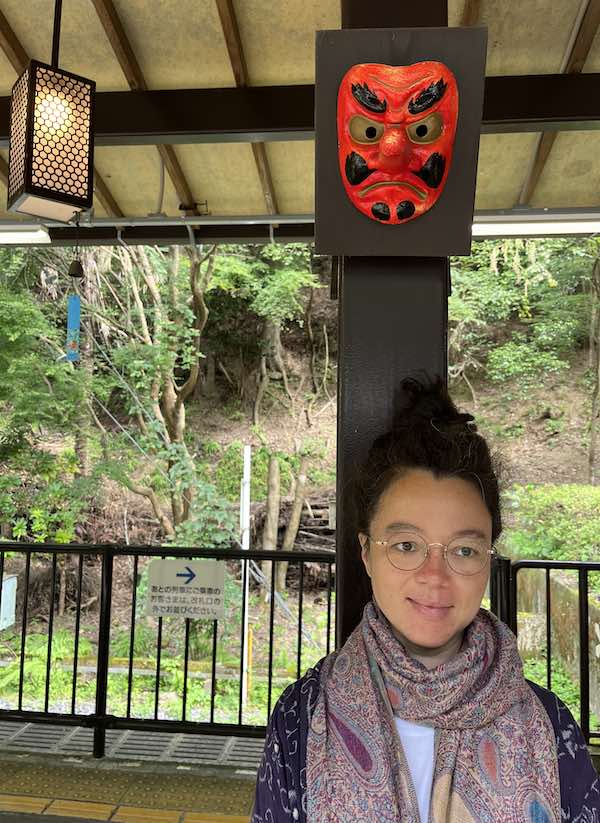 Woman in glasses and scarf stands at a Kyoto mountain station beneath a red traditional mask, linking Gion Matsuri travel with guru yoga and Dharma awakening.