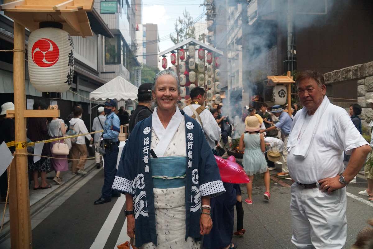 Female Dharma teacher at Kyoto's Gion Matsuri Festival, standing near lanterns, smoke, and a yamaboko float on a guru yoga path.