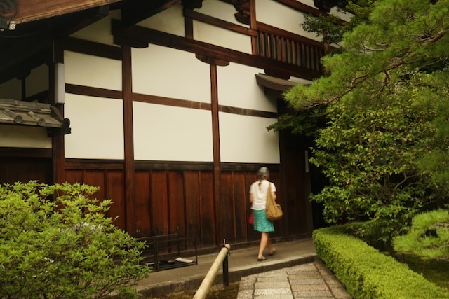 Woman walking beside a traditional Kyoto machiya with garden greenery, evoking Dharma tradition, guru yoga, and the path with a spiritual mentor.