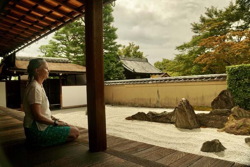 Woman seated on a wooden temple veranda contemplating a Kyoto Zen rock garden, reflecting guru yoga, Dharma tradition, and awakening with a spiritual mentor.