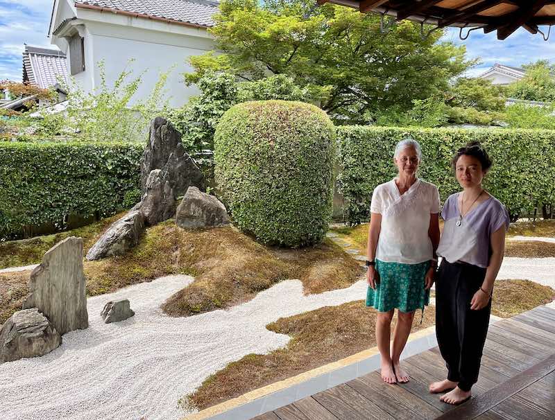 Two women stand barefoot on a wooden veranda beside a Kyoto machiya garden of raked gravel, moss, stones, and hedges, reflecting guru yoga and Dharma mentorship.