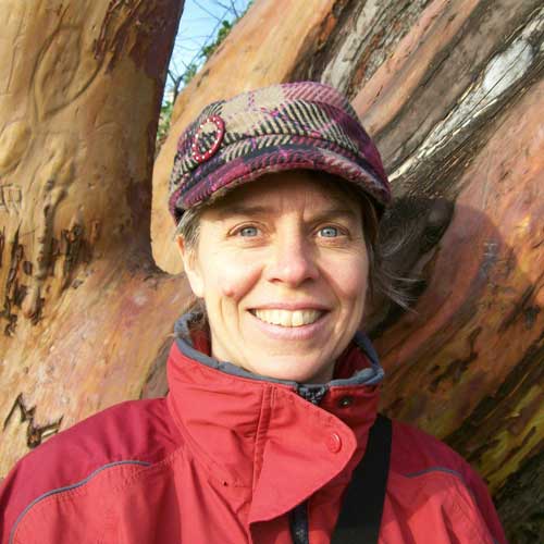 Maureen Smith smiles outdoors in a red jacket and patterned cap beside large tree trunks, illustrating a mighty meditator and Dharma Articles for Awakening mediation retreat story.
