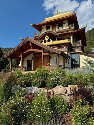 Tibetan-style Buddhist temple with copper-colored roofs, balconies, and garden flowers beneath a blue sky, evoking Dharma practice and aspiration.