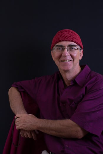 Portrait of a smiling Dharma teacher in a red knit cap and purple shirt against a dark background, for Awakening, community and The Path.