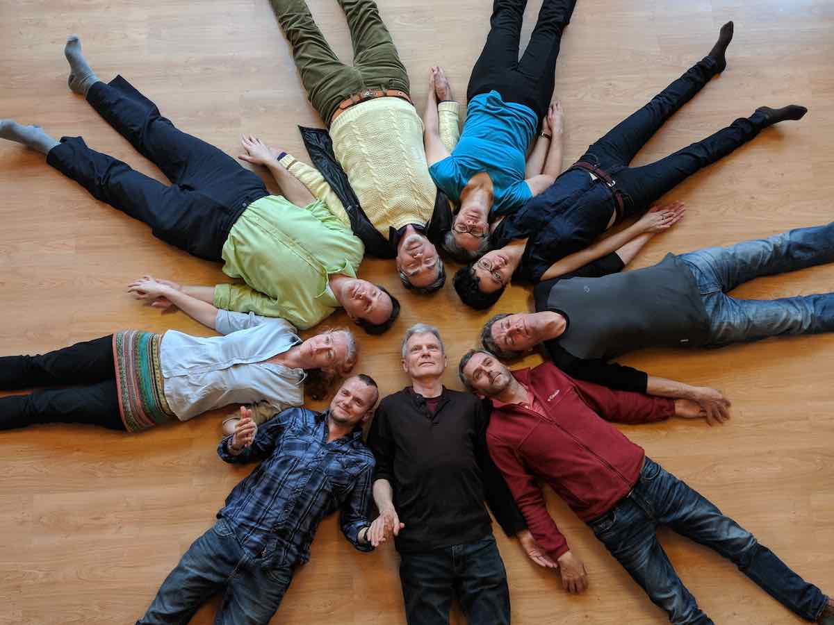 Spiritual community lying in a circle on a wooden floor, heads together in a star shape, suggesting Awakening, shared Dharma practice, and The Path.