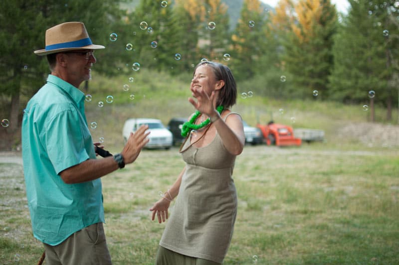Two adults smile and play with bubbles in a grassy outdoor community setting, suggesting Awakening, Dharma practice, and The Path of joyful presence.