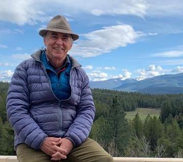 Elder Dharma teacher in a hat and blue jacket seated before forested mountains, evoking Integral Evolution, community, and 21st-Century Awakening.