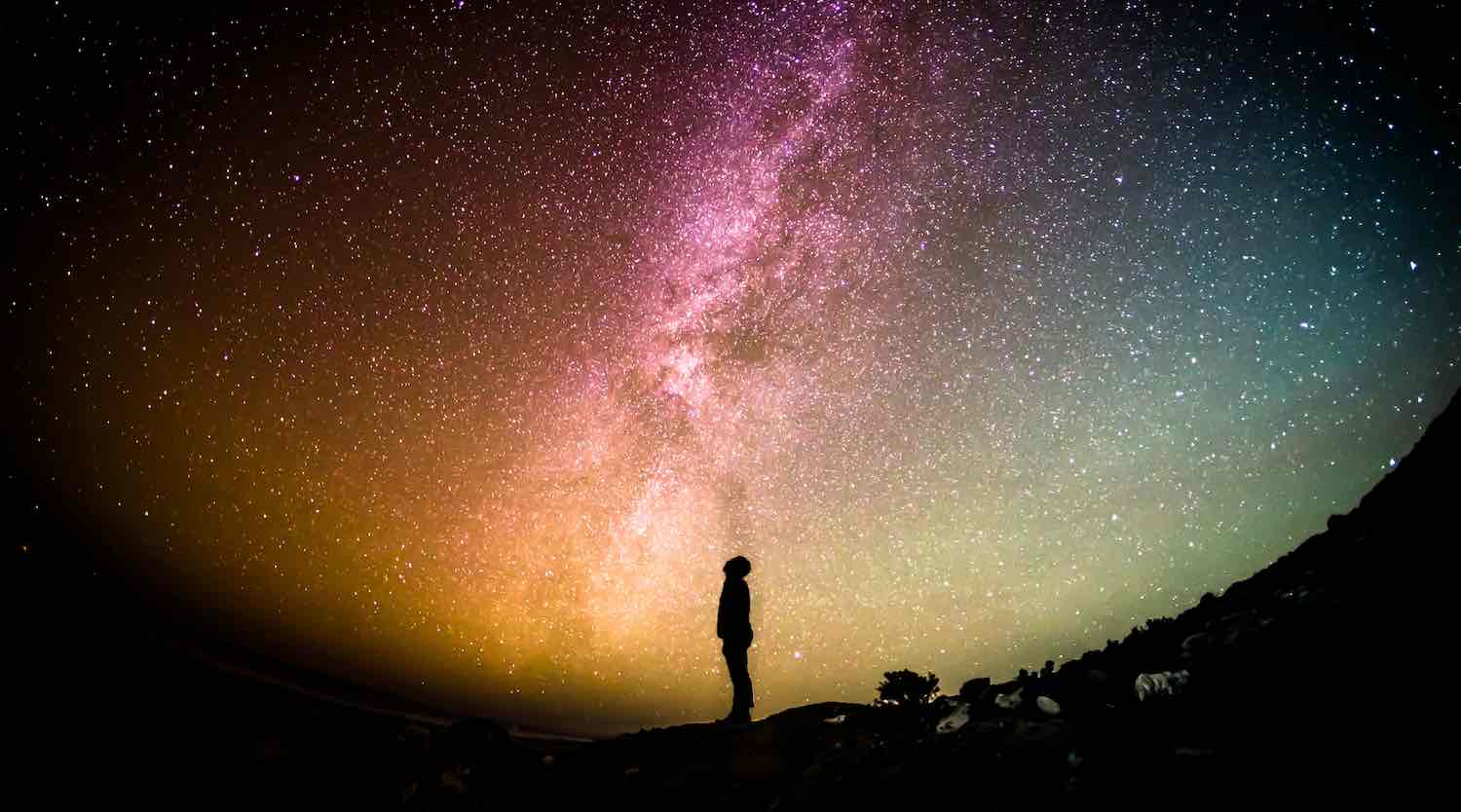 Silhouetted person stands on a rocky ridge beneath a vivid Milky Way sky, evoking Astro-Dharma, Saturn mastery, and contemplative awakening.