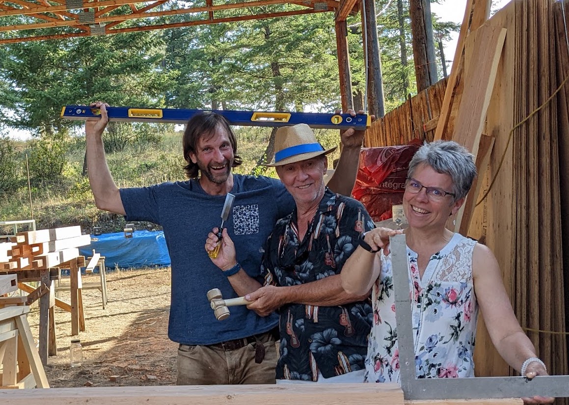 Three Dharma community members smiling at a timber construction site, holding tools and a level as a symbol of conscious collaboration and awakening.