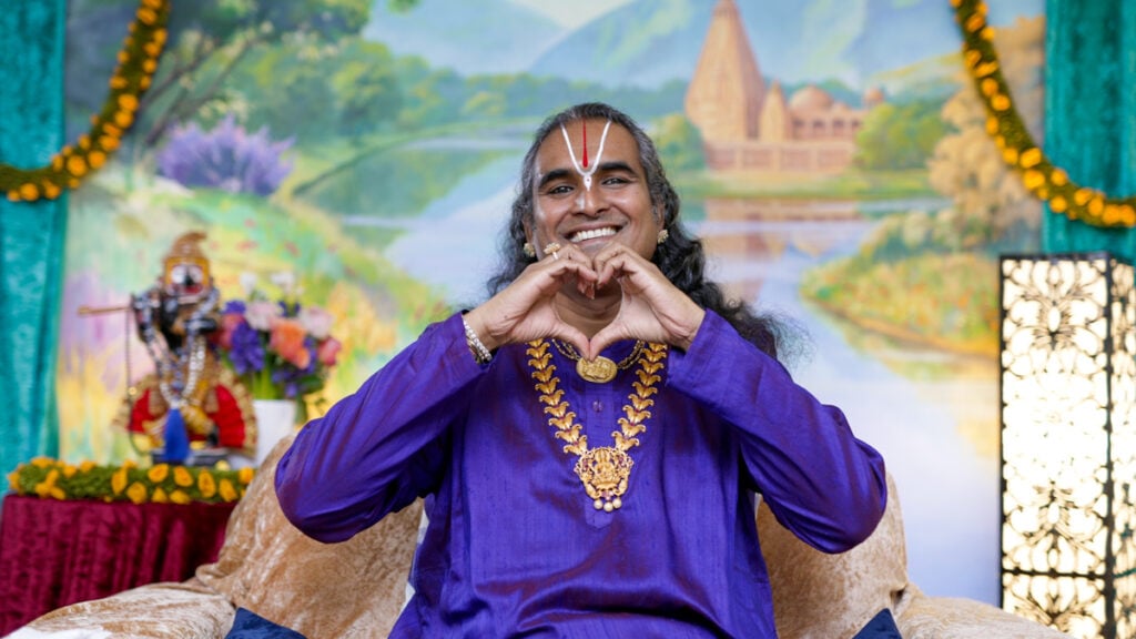 Smiling spiritual teacher in a purple robe forming a heart gesture before a devotional backdrop, evoking bhakti yoga, meditation, and divine love.