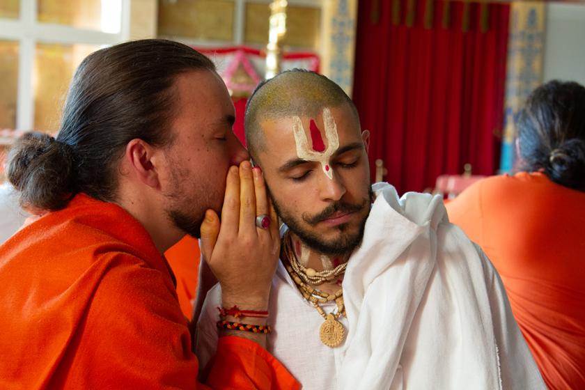 A devotee in orange robes whispers to a seated man with Vaishnava tilak and prayer beads, reflecting bhakti lifestyle and spiritual guidance.