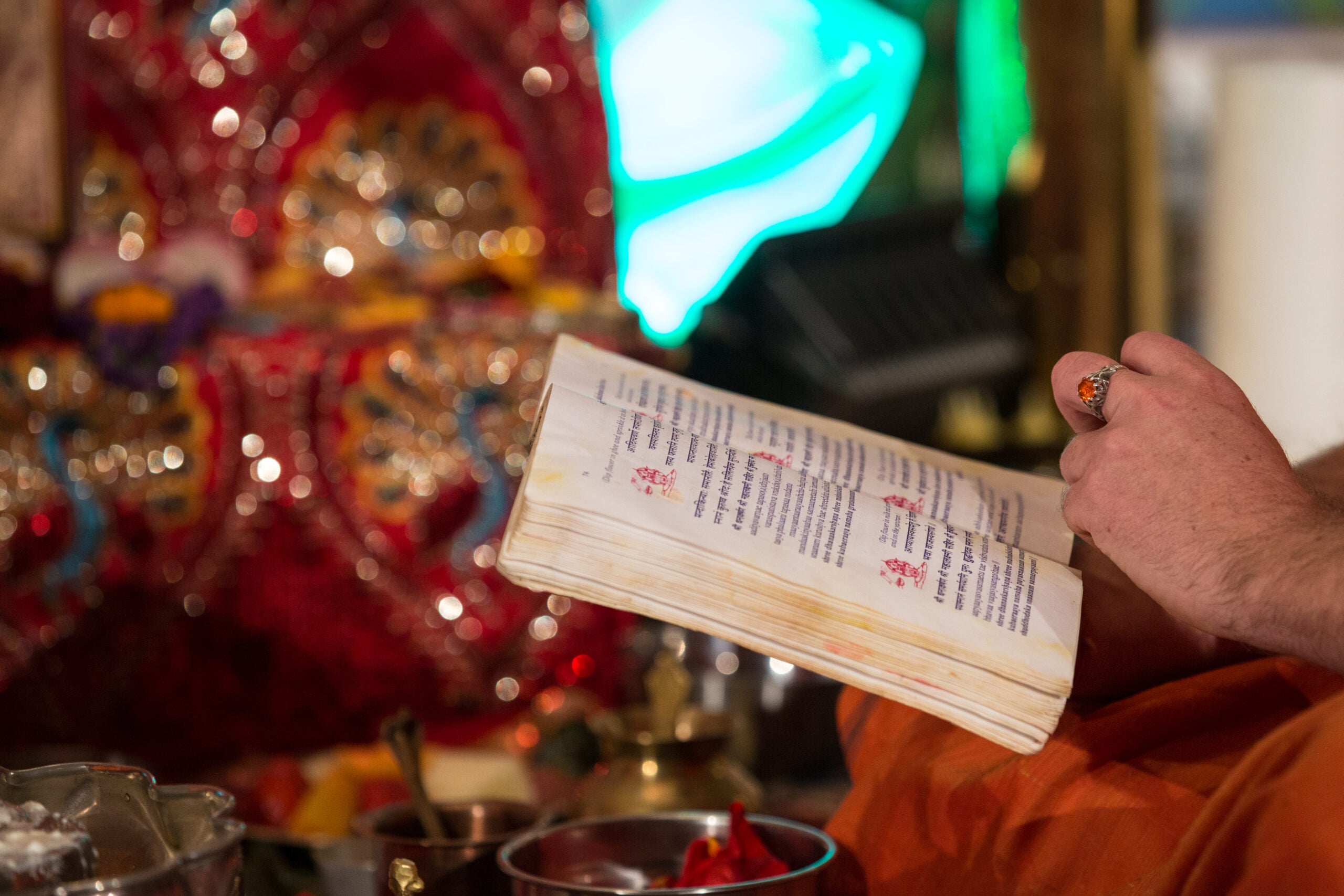 A devotee holds an open Hindu prayer book before a brightly decorated altar, suggesting Knowledge, Vedanta, and reflection on the personal and impersonal God.