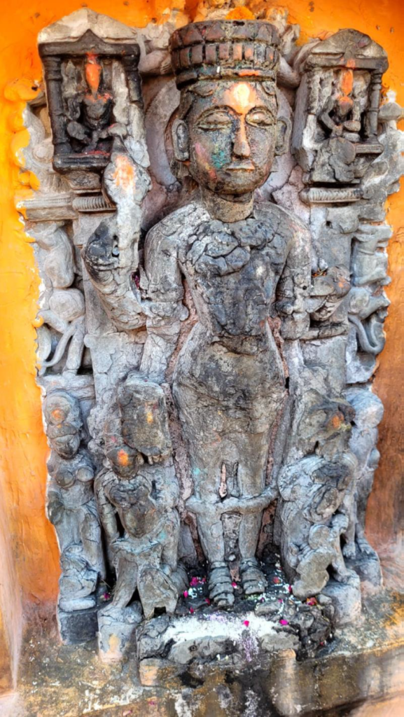 Weathered stone idol, likely Vishnu, set in an orange niche at Prithvinath Temple in Gonda, showing eroded carvings and offerings below.