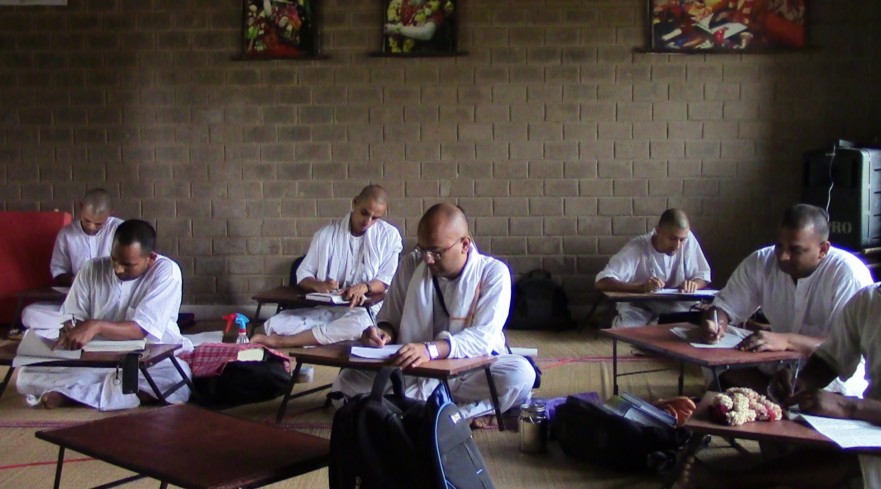 Spiritual students in white robes sit on floor mats writing at low desks in a simple classroom, reflecting guidance, autonomy, and learning.
