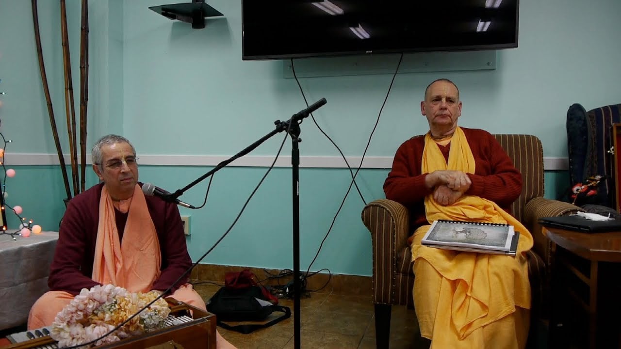 Two ISKCON swamis in saffron and maroon robes sit in a temple room with microphones during a talk remembering Dhirasanta Dasa Goswami.