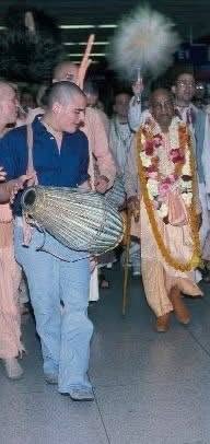 ISKCON devotees in a kirtan procession, with a man playing mridanga beside a garlanded devotee, honoring Tranakarta Das ACBSP.