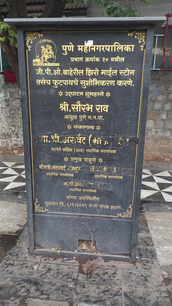Damaged black stone plaque with Marathi text at Pune’s Zero Stone site, showing scratches, dirt and neglected civic surroundings.