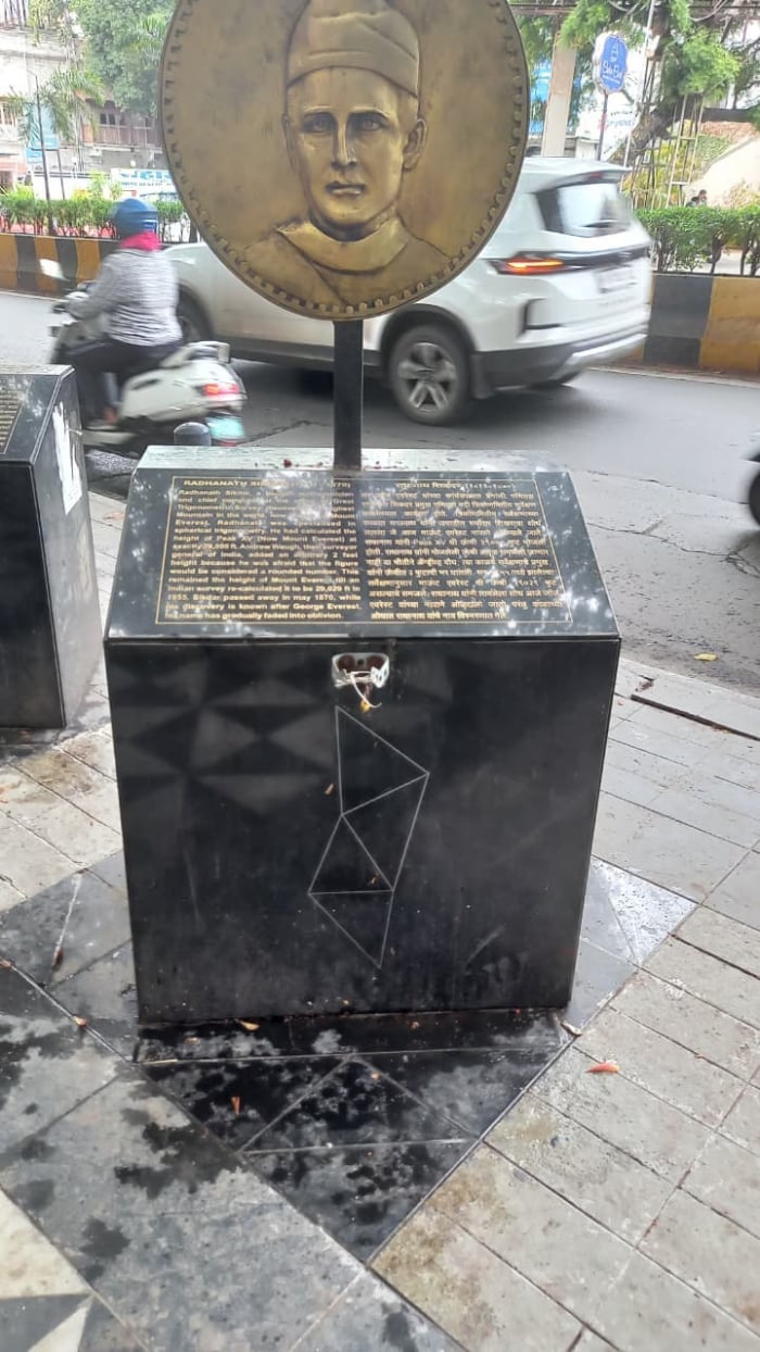 Pune Zero Stone memorial with a bronze portrait plaque on a black pedestal, showing scratched vandalism and neglect beside a busy city road.