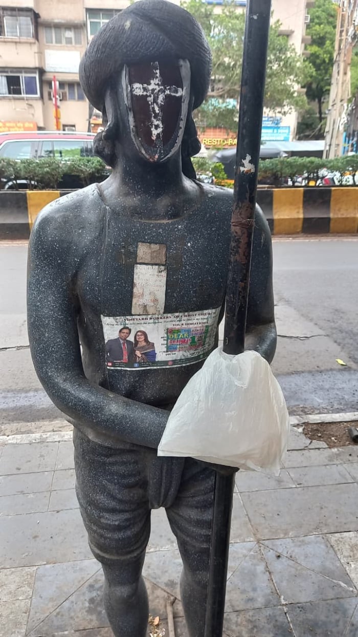 Vandalised black statue near Pune’s historic Zero Stone, with defaced face, pasted posters and a plastic bag tied to a pole on a city footpath.