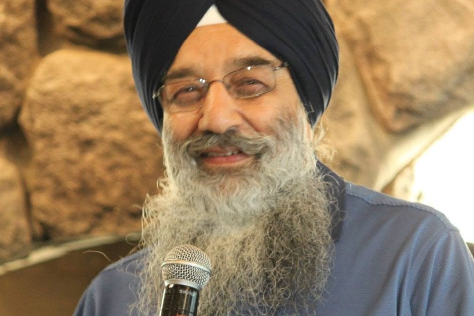 S. Kuldeep Singh Ji, a Sikh elder with a navy turban, glasses, and long white beard, smiling while holding a microphone at a gathering.
