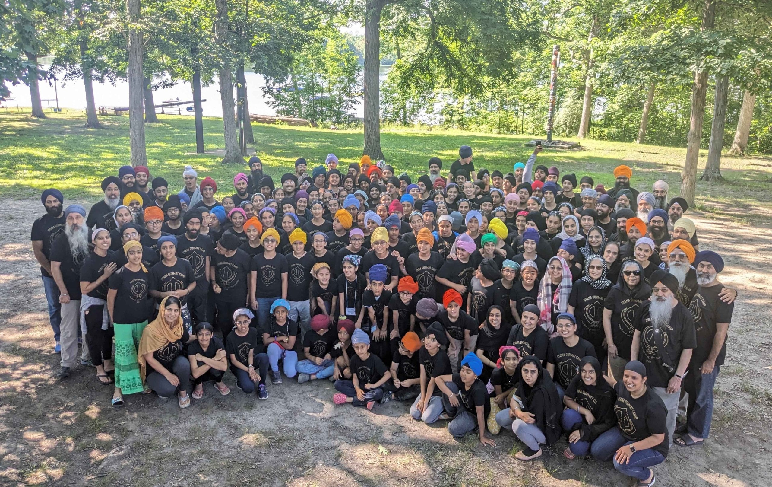Large Sikh Gurmat camp group photo outdoors, with youth and volunteers in matching shirts and colorful turbans gathered near trees and a lake.