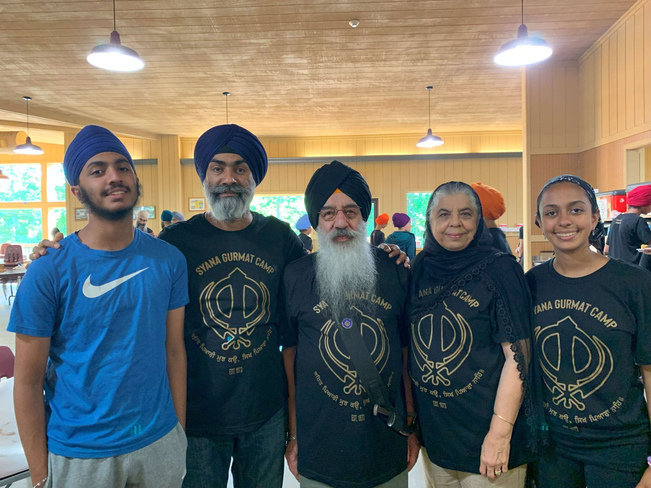 Five Sikh community members, including elders in turbans and Syana Gurmat Camp shirts, pose together indoors in remembrance of S. Kuldeep Singh Ji.