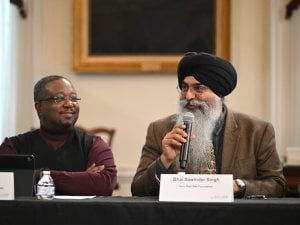 Sikh panelist in a black turban speaking into a microphone at a table, related to remembering S. Kuldeep Singh Ji and Gurmat Camps