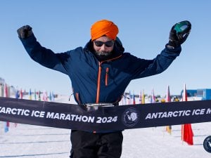 Sikh runner in an orange turban raises his arms at the Antarctic Ice Marathon 2024 finish line on a bright snowy course.