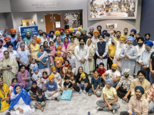 Large Sikh community group gathered indoors at a gurdwara or camp hall, with children, adults, and elders posing together for a Gurmat Camp remembrance.