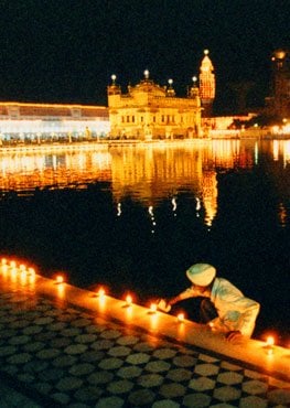 Person lighting rows of diyas beside the sarovar at Sri Harmandir Sahib, the Golden Temple, at night with glowing reflections on the water.