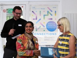 A smiling woman receives a blue book or award at a community event, with a speaker holding a microphone and a Connections poster behind them.
