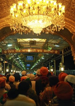 Sikh congregation seated beneath a glowing chandelier inside a gurdwara, reflecting SikhNet community, faith, youth creativity and Sikh culture.
