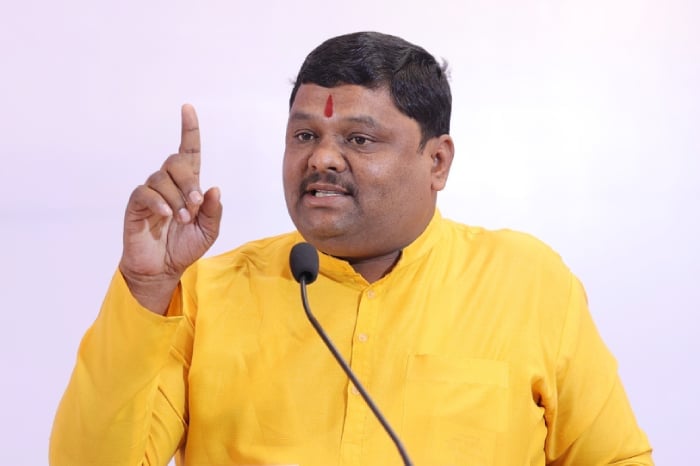A man in a yellow kurta with a red tilak speaks at a microphone, raising one finger, linked to the Shri Tuljabhavani Mandir land scam inquiry.