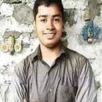Smiling adult man in a grey shirt before a textured wall, accompanying a Sri Rangam travelogue on Srirangam and Sri Ranganathaswamy.