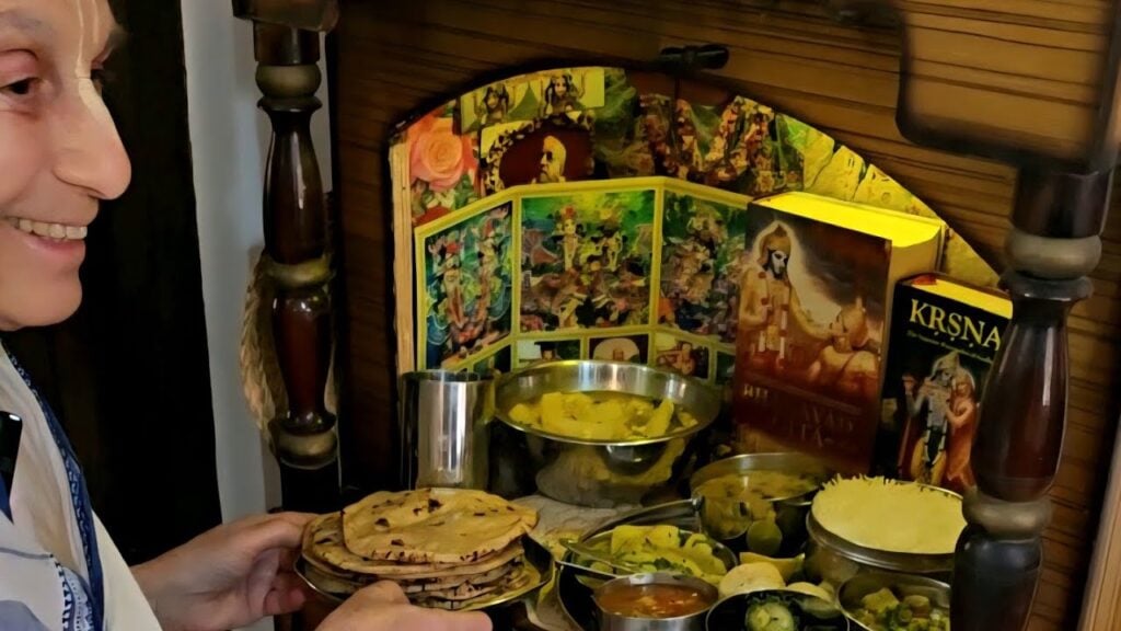 Daivisakti Mataji holds chapatis beside a devotional lunch offering for Srila Prabhupada, with Krishna books, bowls, rice and vegetables.