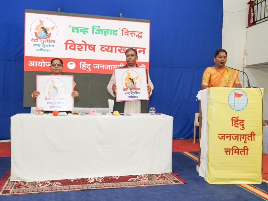 Speakers at a Satara Hindu Janajagruti Samiti symposium display Beti Surakshit campaign posters as a woman addresses attendees from a podium.