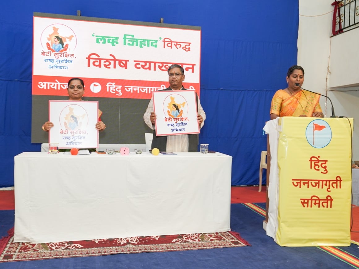 Speakers at a Satara Hindu Janajagruti Samiti symposium display Beti Surakshit campaign posters as a woman addresses attendees from a podium.