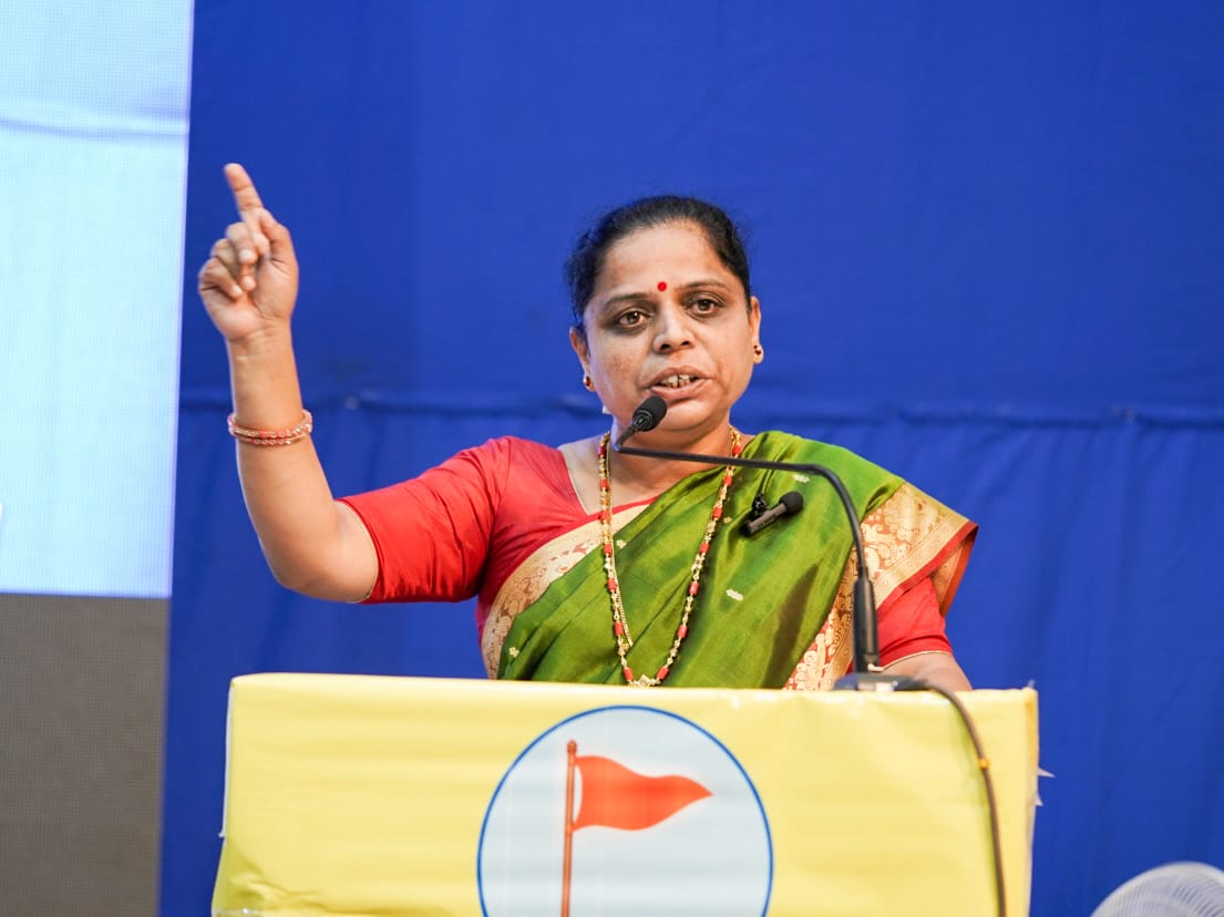 Woman speaker in a green sari addresses a Satara symposium from a podium, raising her finger during a Hindu Janajagruti Samiti public event.