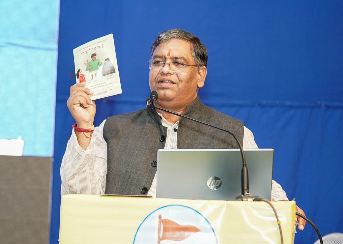 Speaker at a Satara Hindu Janajagruti Samiti symposium holds a booklet beside a laptop and microphone during a Corporate Jihad awareness event.